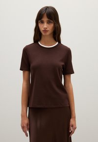 T-shirt marrone a maniche corte con colletto a costine bianche, realizzato in un tessuto liscio. La vestibilità è rilassata e presenta un orlo dritto.