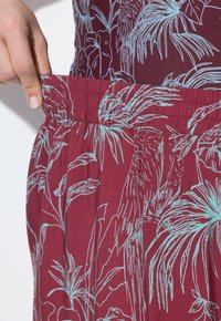 Mörkröd tyg med ett blått tropiskt blad- och fågelmönster. Har en elastisk midja och mjukt, texturerat material. Närbild.