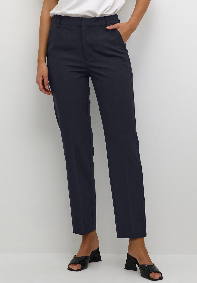 Femme portant un pantalon droit bleu marine avec poches latérales et des sandales à talons blocs ouvertes noires, debout devant un fond blanc.