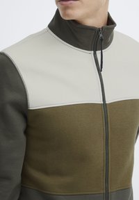 Giacca a zip in fleece con colletto alto, in sezioni beige e verde oliva, con cuciture visibili e chiusura con zip.