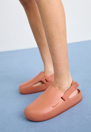 Person iført laksefarvede Nike slip-on træsko med justerbare hælremme på en blå gulv.