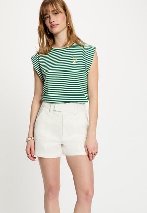 Donna che indossa una canottiera a righe verdi e bianche con un piccolo emblema di aragosta, pantaloncini bianchi con vita alta e gioielli dorati.