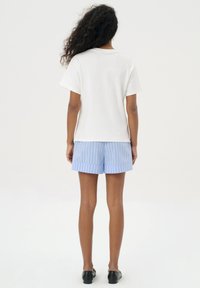 Biała bawełniana koszulka zestawiona z jasnoniebieskimi paskowanymi szortami, o luźnym kroju i gładkim przodzie. Całość dopełniają czarne buty wsuwane.