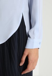 Ljusblå långärmad blus med rundad nederkant och knäppte manschetter, i kombination med marinblå randiga byxor. Slät tygkvalitet, skräddarsydd passform.