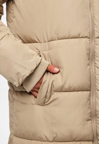 Beige pufferjakke med teksturert overflate, en frontlomme og ribbestrikkede mansjetter. Glidelåslukking foran.