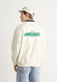 Felpa crema con grafica verde sul retro che presenta un design di montagna e testo. Materiale in cotone morbido, vestibilità rilassata, polsini e orlo a coste.