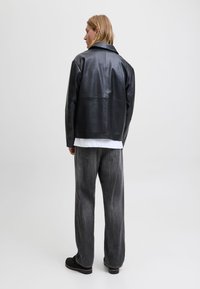 Melna ādas jaka ar strukturētu dizainu, platu apkakli un redzamām šuvju detaļām, kombinēta ar pelēnām plaša piegriezuma džinsiem un melniem apaviem.