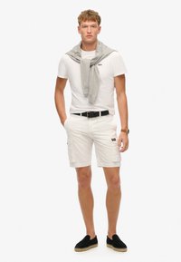 Pantalones cortos de carga blancos con bolsillos laterales, combinados con una camiseta blanca ajustada. Un suéter gris claro caído sobre los hombros. Zapatos negros tipo slip-on.