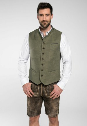 Grüne Wollweste mit hohem Kragen, Fronttaschen und braunen Knöpfen, kombiniert mit braunen Lederhosen mit dekorativer Stickerei.