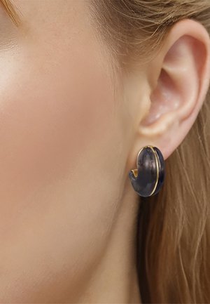 Nærbillede af en persons øre med en buet sort og gylden ørering i hoop-stil, hvor kort, lysebrunt hår er synligt.