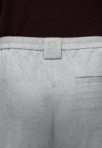 Gros plan sur un pantalon gris clair à motif chevrons avec une étiquette à la taille comportant le logo "MM" et une poche arrière sur le côté droit.