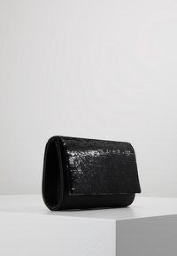 Svart kuvertväska med glittrande paljetter på klaffen, rektangulär form, placerad på en vit yta med en neutral grå bakgrund.