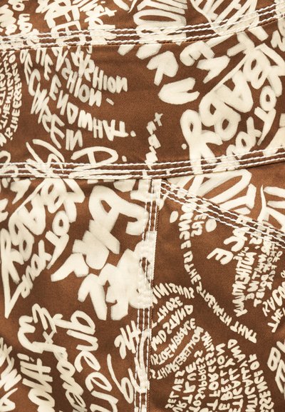 Καφέ ύφασμα με λευκό κείμενο σε στυλ γκράφιτι και εμφανείς ραφές που σχηματίζουν γεωμετρικά σχέδια στο υλικό.