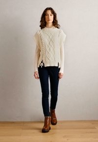 Pull sans manches en tricot crème avec motif en torsades, associé à un jean foncé et des bottines marron, avec des liens sur les côtés pour un ajustement réglable.