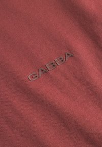 Mørkerødt stof med fin tekstur, der har det prægede logo "GABBA" i en subtil, mørkere nuance. Fokus på blødhed og minimalistisk design.