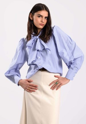 Blusa a rayas azul claro con mangas abullonadas y un gran lazo, combinada con una falda de color crema. Detalles notables de botones.