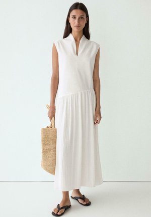 Donna con vestito midi bianco senza maniche che tiene una borsa a tracolla intrecciata, indossa sandali neri, in piedi su uno sfondo chiaro uniforme.