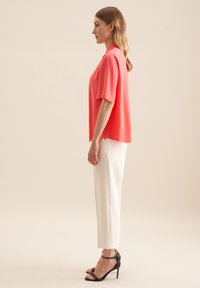 Blusa corallo a maniche corte con una vestibilità ampia, abbinata a pantaloni bianchi su misura e sandali neri con tacco alto. Tessuto morbido e design elegante.