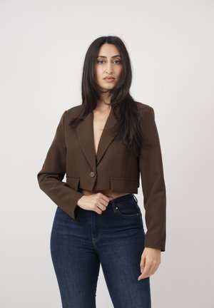 Frau mit langen dunklen Haaren, die ein kurzes braunes Blazer und dunkelblaue Jeans trägt, vor einem schlichten hellgrauen Hintergrund stehend.