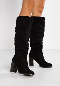 Botas negras sobre la rodilla hechas de suave gamuza con un tacón ancho, que presentan un diseño arrugado y una textura lisa.