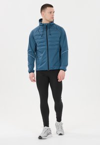 Chaqueta con capucha para hombre en color azul verdoso, con cremallera frontal, diseño acolchado y costuras en contraste. Combinada con leggins negros y zapatillas de correr.