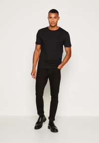 Svart bomullströja med korta ärmar och rund halsringning, tillsammans med svarta slim-fit jeans och svarta snörskor, mot en enfärgad bakgrund.