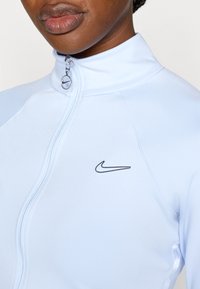 Svetlo modra jopa z zadrgo iz gladkega materiala, z visokim ovratnikom in črnim logotipom Nike na prsih. Opremljena s srebrno zadrgo.