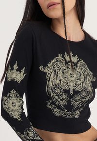 Zwarte ribgebreide crop top met gouden grafische ontwerpen, waaronder kronen en bloempatronen op de mouwen en in het midden.