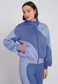 Giacca leggera con zip in diverse tonalità di blu, con una superficie liscia, dotata di un alto colletto e tasche laterali.