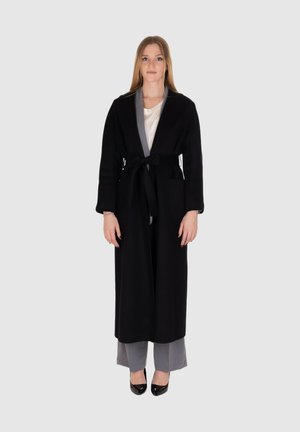 Lungo cappotto nero con cintura, tasche laterali e interno grigio. Indossato sopra pantaloni larghi grigi, abbinato a tacchi neri.