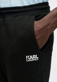 Roce rokā melnu sporta biksu kabatā ar baltu KARL LAGERFELD logo uz augšstilba, valkājot gaismas zila krekla.