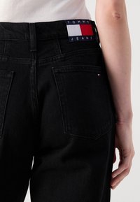 Personne portant un jean noir Tommy avec des écussons logo visibles à la ceinture arrière et à la poche, une chemise blanche et un bras avec une cicatrice au coude.
