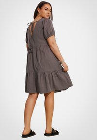 Vestido gris hasta las rodillas con mangas cortas abullonadas, detalle de lazo en la espalda y dobladillo con volantes. Combinado con sandalias negras tipo slide con adornos.