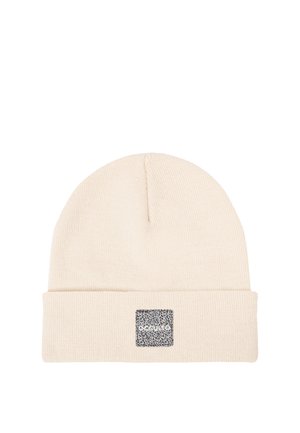 Mann mit leichtem Schnurrbart, der eine beige Mütze und ein schwarzes kariertes Hemd trägt, posiert mit den Händen hinter dem Kopf vor einem einfarbigen weißen Hintergrund.