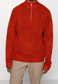 Strickjacke mit Reißverschluss in leuchtendem Orange, hochgeschlossen, mit strukturiertem Stoff und gerippten Bündchen und Saum, kombiniert mit beigen Hosen.