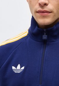 Mann trägt eine marineblaue Adidas-Trainingsjacke mit Frontreißverschluss und gelben Streifen auf der Schulter, die das Adidas-Logo zeigt.
