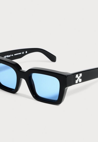 Lunettes de soleil rectangulaires noires avec des verres bleu clair et un logo croix blanc sur la branche, sur un fond gris clair.