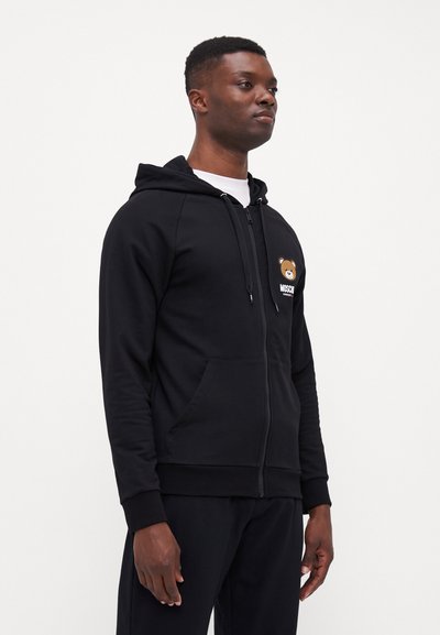 Black zip-up hoodie made of soft fabric, featuring a front pocket and a bear graphic on the left chest. Fitted cuffs and hood.