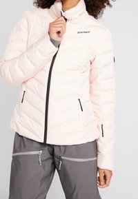 Růžová zateplená bunda s vysokým límcem, předním zipem a vertikálním prošitím. Má boční kapsy a logo na prsou.