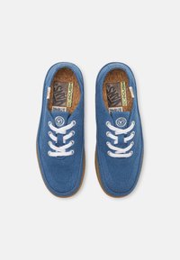 Os sapatos de lona azuis apresentam atacadores brancos, um superior texturizado e uma palmilha de cortiça. A sola é de borracha castanha, proporcionando um contraste com o azul.