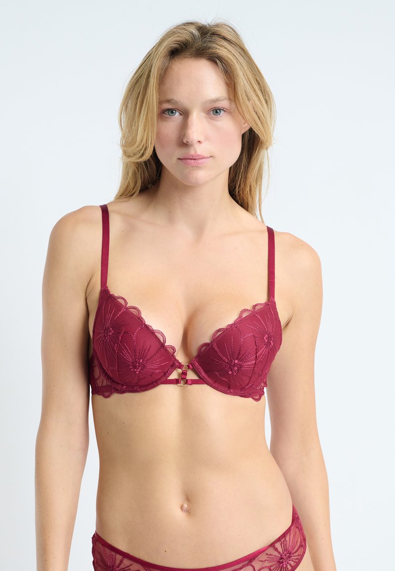 Sostén de encaje burdeos con bordado floral, bordes festoneados, copas acolchadas y tirantes delgados ajustables. Presenta un detalle decorativo en la parte frontal.
