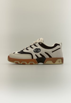 Basket basse blanche et noire avec semelle marron, lacets et logo DC sur le côté, présentée sur un fond clair uni.