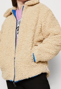 Béžová huňatá bunda s texturovaným povrchem, modrou podšívkou a zipovým zapínáním. Obsahuje límec a boční kapsy.