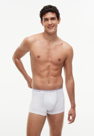 Ung mand med mørkt krøllet hår der smiler, stående uden skjorte, iført hvide Tommy Hilfiger boxershorts mod en hvid baggrund.