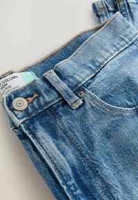Dámské modré džíny s vysokým pasem, s jemným ombré efektem, viditelným švem a stříbrným knoflíkem. Na pásku je připojena etiketa s velikostí a pokyny k údržbě.