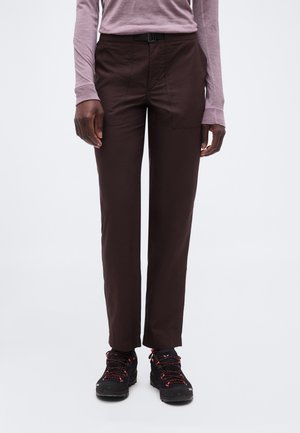 Személy, aki sötétbarna egyenes szárú nadrágot, fekete túracipőket piros fűzőkkel, és hosszú ujjú levendulakék felsőt visel egyszínű háttér előtt.