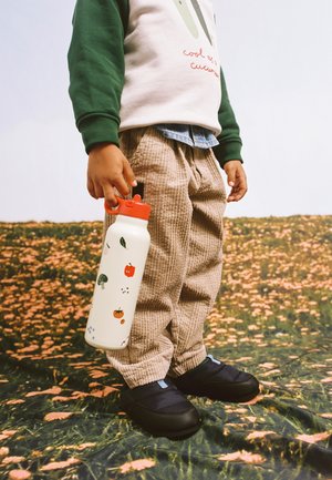 Barn iført beige fløjlsbukser, grøn og hvid sweatshirt, der holder en hvid vandflaske med frugtillustrationer, stående på blomsterjord.