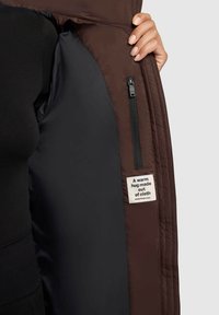 Veste marron avec un intérieur noir, comportant une poche zippée et une étiquette tissée qui indique : "Un câlin chaud fait de tissu."
