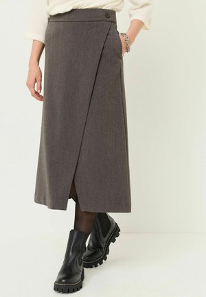 Personne portant une jupe portefeuille gris foncé, des collants noirs, des bottines noires épaisses et un pull blanc avec une main dans la poche de la jupe.