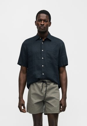 Egy férfi tengerészkék, rövid ujjú, gombos ingben és világosszürke, zsinóros rövidnadrágban áll sima háttér előtt.